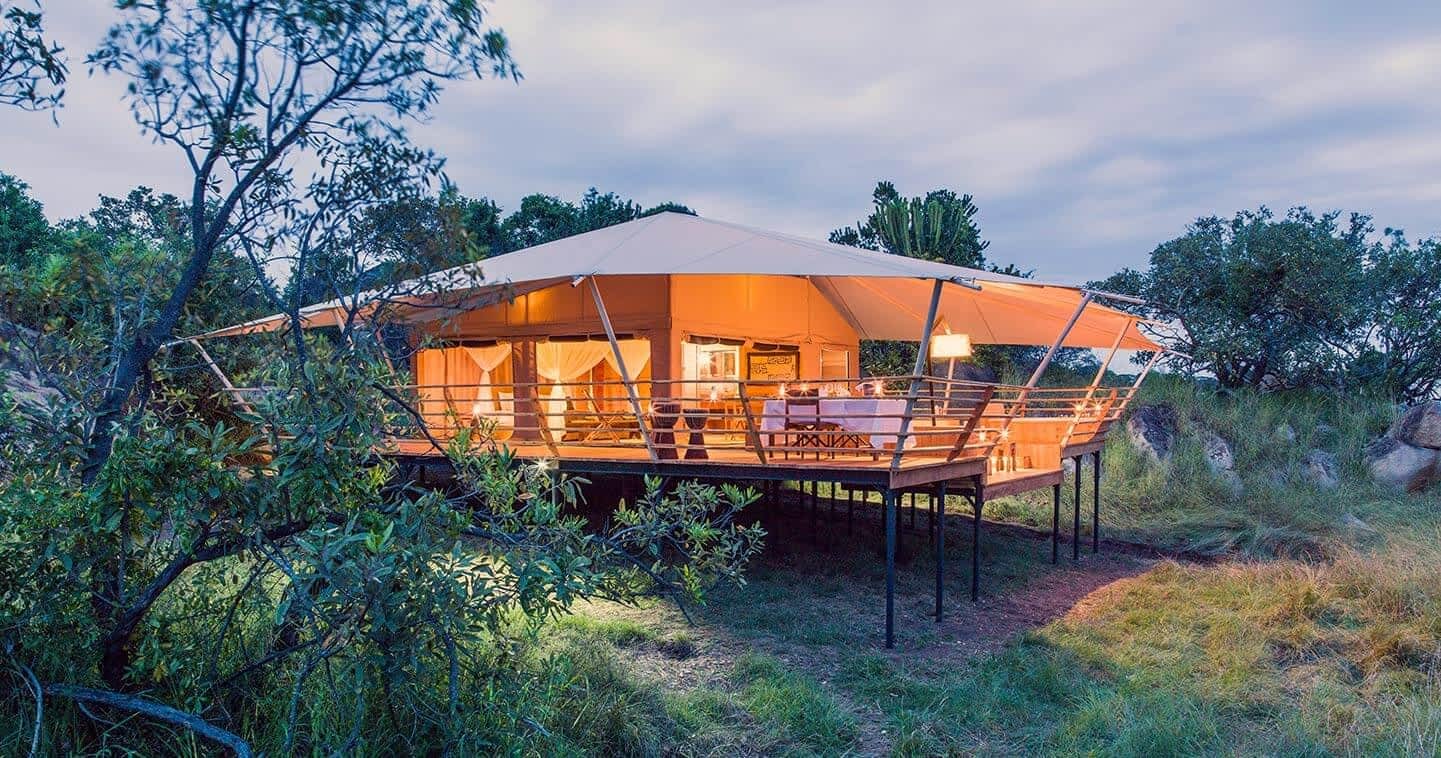 A high-end luxury tented camp interior featuring fine linen bedding, polished wood furniture, and local African textiles. The tent opens up to a private deck with an expansive view of the golden savanna. The lighting is warm and candlelit, creating an intimate, romantic, and extremely comfortable sanctuary in the heart of the wild. Every detail speaks to sophisticated grace and handcrafted quality.