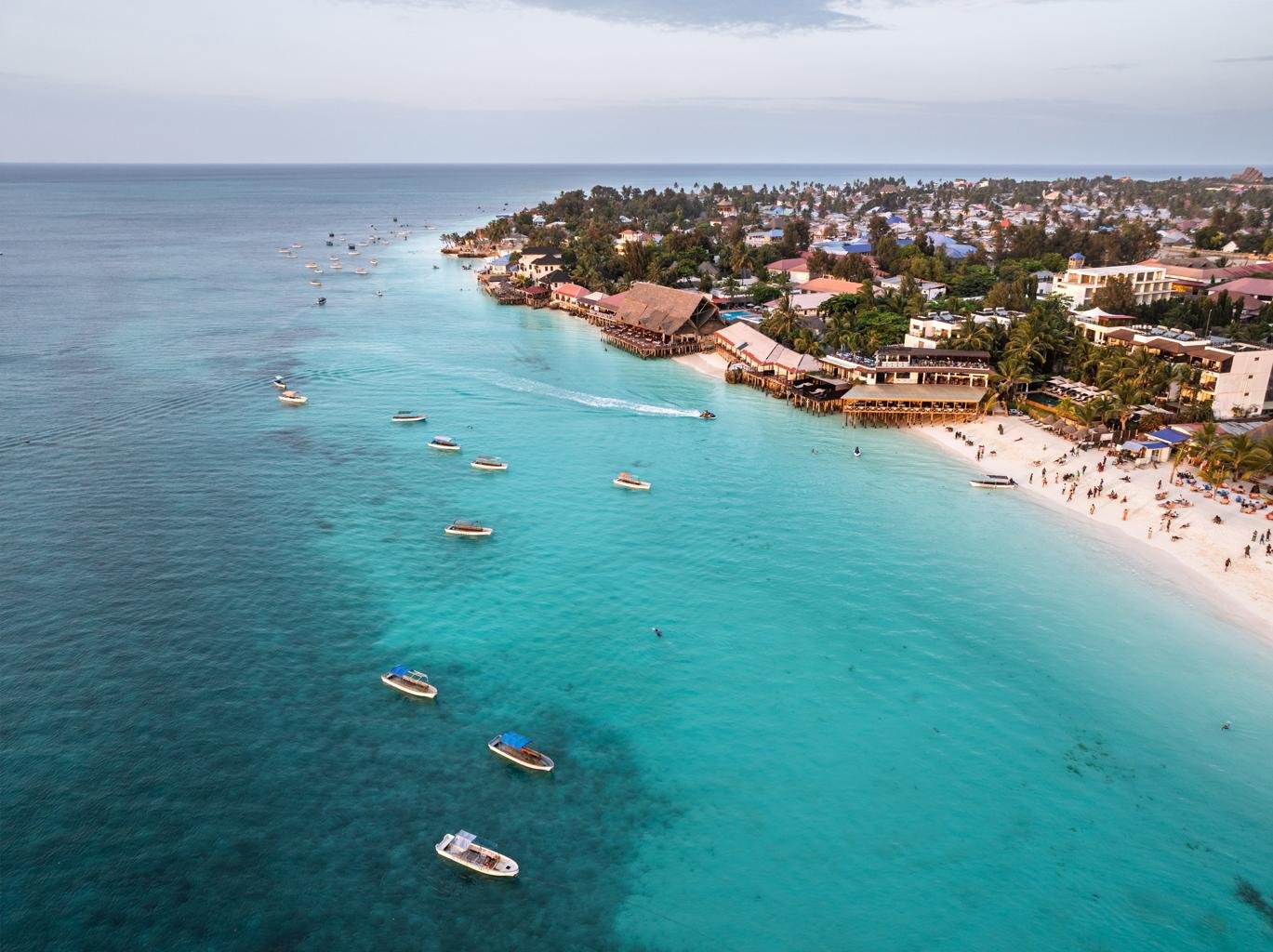 Crystal clear turquoise waters and white sandy beaches of Zanzibar at sunset. Dhow boats are silhouetted against a vibrant orange and pink sky. A peaceful, luxurious tropical vibe that balances the wildness of the mainland safari.