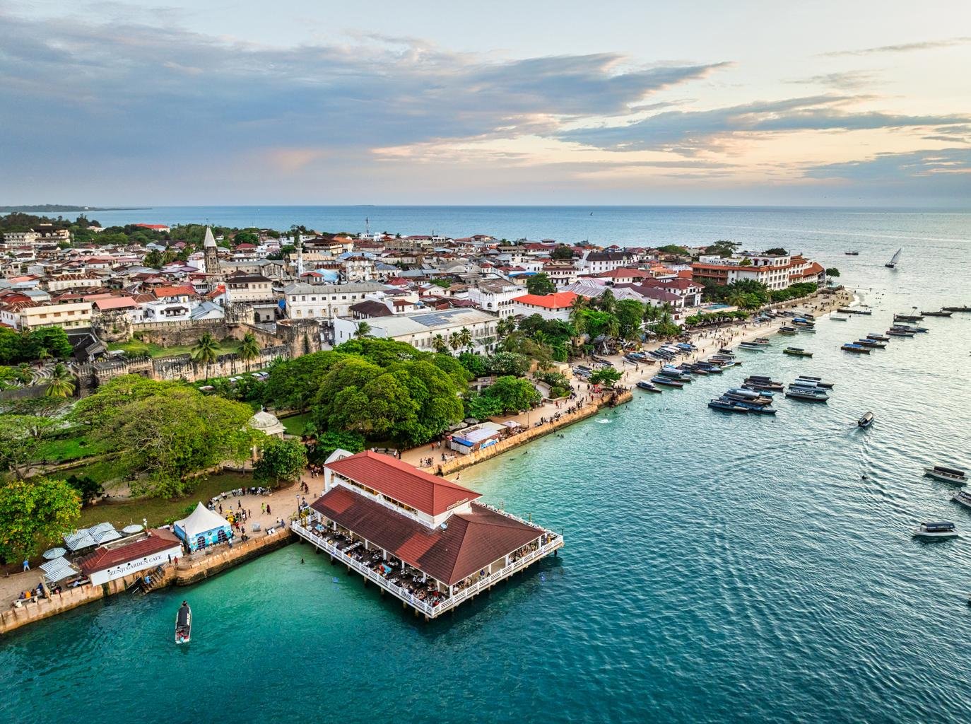 The turquoise waters of Zanzibar crashing against white sandy beaches. A traditional dhow sailboat is anchored in the shallow water. The lighting is bright midday sun, emphasizing the vibrant colors of the Indian Ocean. This image represents the relaxing, luxury end to a Tanzanian safari, styled with high-end resort photography in mind.